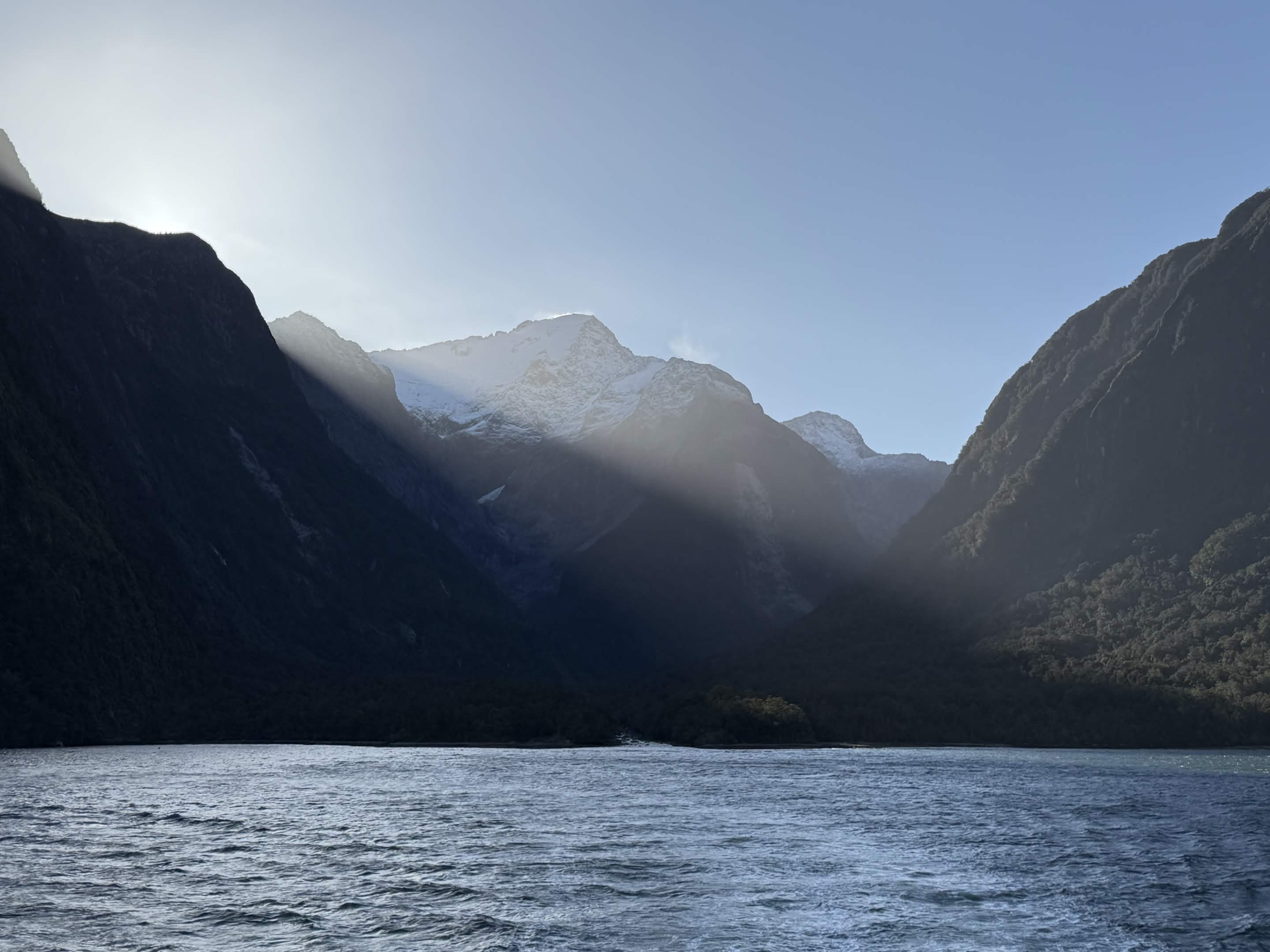 Sun cresting a snow-dusted peak at Milford Sound, rays raking across dark forested cliffs down into the fiord